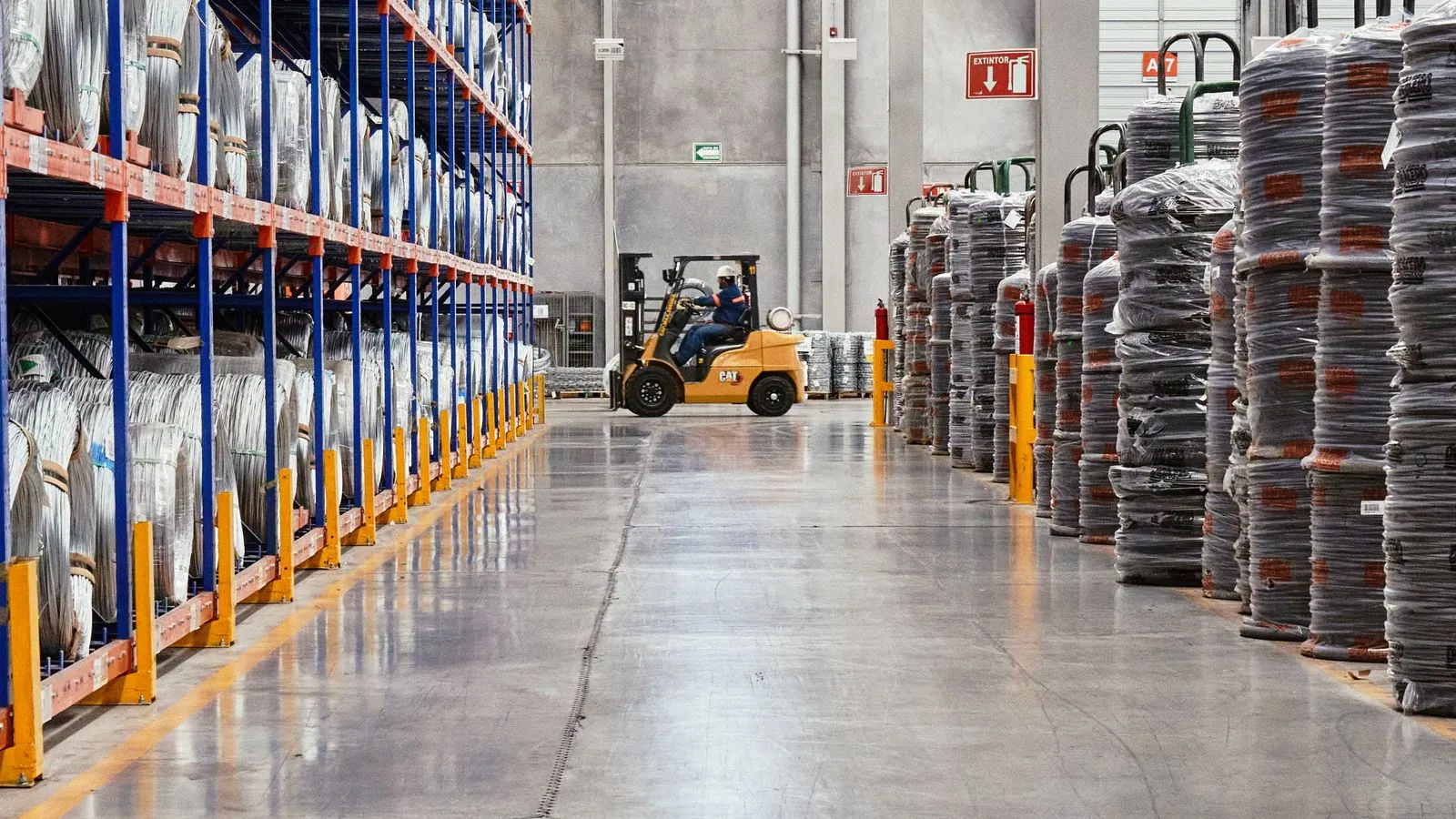 Bodega industrial con estanter&iacute;as met&aacute;licas cargadas de equipos embalados y un montacargas circulando entre los pasillos